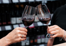 Zwei Personen stoßen mit Rotweingläsern an, Weinregale im Hintergrund.