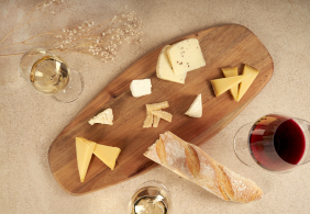 Holzbrett mit verschiedenen Käsesorten, Baguette und Weingläsern auf heller Oberfläche.