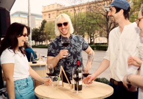 Vier Personen lachen und genießen Rotwein an einem Stehtisch im Freien.