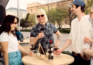 Vier Personen lachen und genießen Rotwein an einem Stehtisch im Freien.