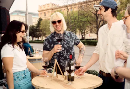 Vier Personen lachen und genießen Rotwein an einem Stehtisch im Freien.