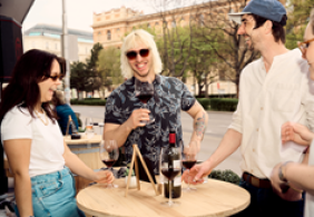 Gruppe von Freunden lacht und trinkt Wein an einem Holzstehtisch im Freien.