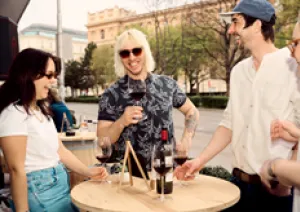 Gruppe von Freunden lacht und trinkt Wein an einem Holzstehtisch im Freien.