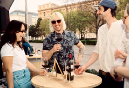 Gruppe von Freunden lacht und trinkt Wein an einem Holzstehtisch im Freien.
