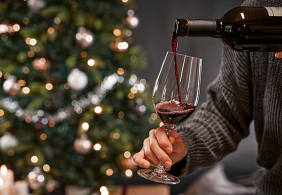 Person schenkt Rotwein in ein Glas neben einem geschmückten Weihnachtsbaum ein.
