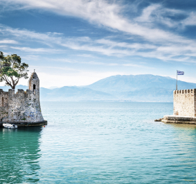Küstenburg mit Baum, klarem Wasser und Bergkulisse unter bewölktem Himmel.