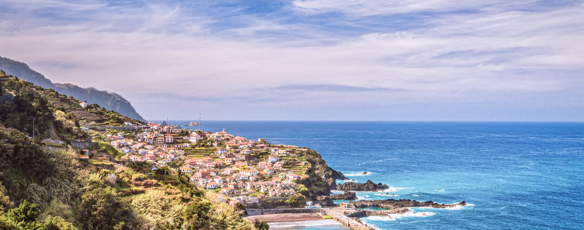 Küstenpanorama mit Dorf auf Hügeln, umgeben von blauem Meer und bewölktem Himmel.