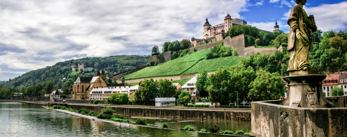 Flusslandschaft mit Burg, Weinbergen und Statue bei bewölktem Himmel.