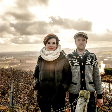 Zwei Personen in Winterkleidung vor bewölkter Landschaft und Weinbergen.