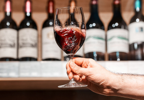 Hand hält ein halbes Glas Rotwein vor einem Weinregal.