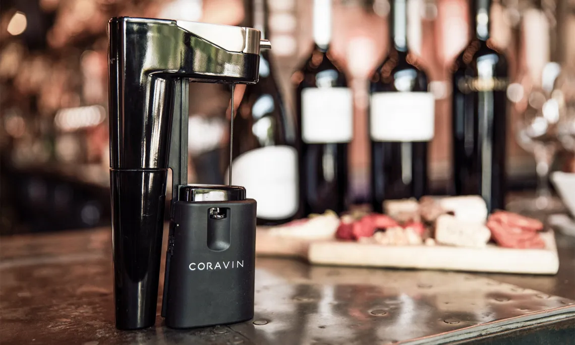 Weinausgießgerät vor unscharfem Hintergrund mit Weinflaschen und Käseplatte.