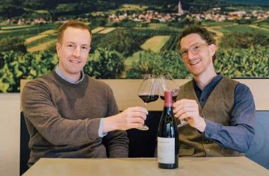 Zwei Männer stoßen mit Rotweingläsern an, Weinflasche auf Holztisch.