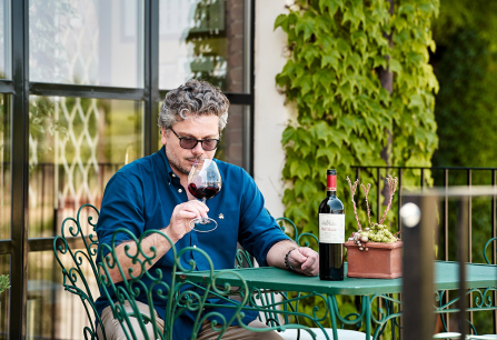 Mann in blauem Hemd genießt Rotwein auf grüner Terrasse mit Pflanzen.