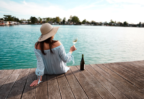 Frau auf Steg mit Hut, hält Weinglas, blickt auf See.
