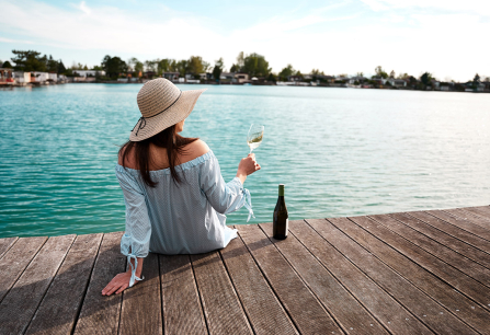 Frau auf Steg mit Hut, hält Weinglas, blickt auf See.