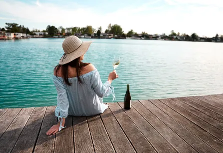 Frau auf Steg mit Hut, hält Weinglas, blickt auf See.