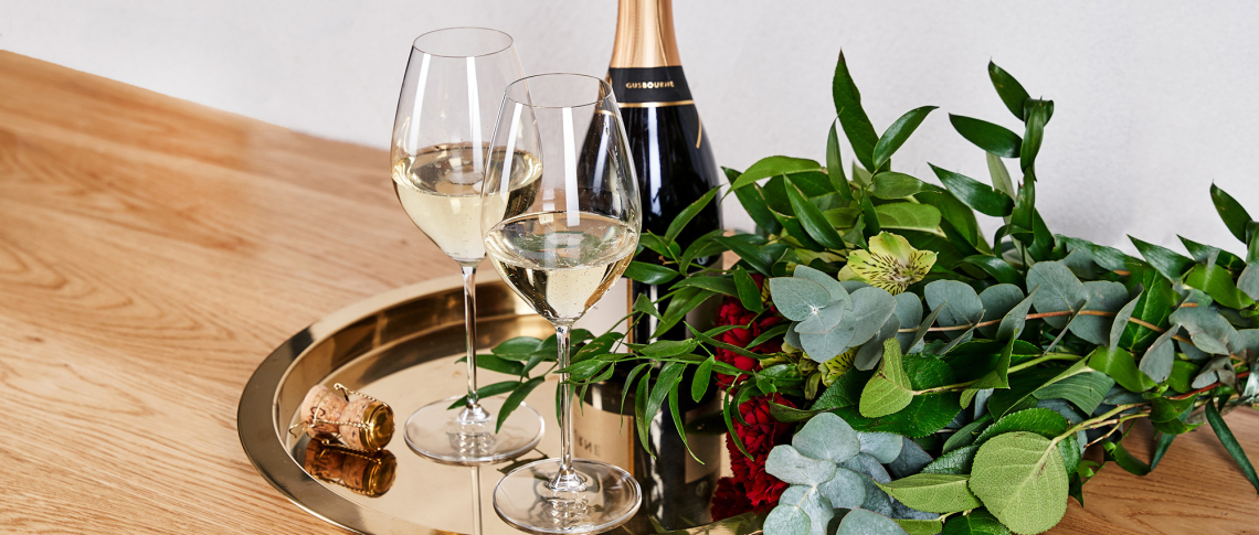 Goldenes Tablett mit Champagnerflasche, zwei Gläsern und Blumenstrauß auf Holztisch.