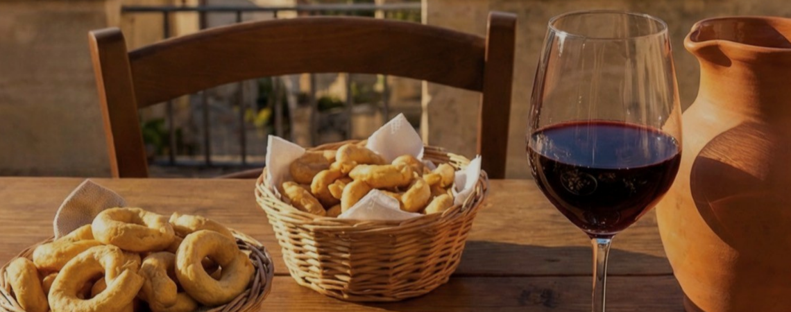 Korb mit Gebäck, Glas Rotwein und Tonkrug auf Holztisch im Freien.