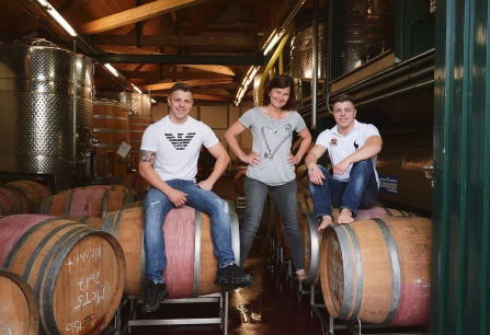 Drei Personen posieren auf Weinfässern in einem Weinkeller.