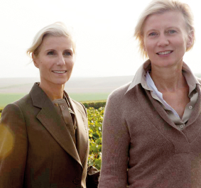 Zwei Frauen in eleganter Kleidung vor einer weitläufigen Landschaft mit Weinreben.