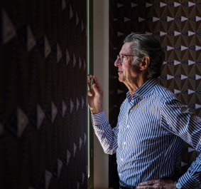 Mann im gestreiften Hemd blickt aus einem Fenster, umgeben von gemusterten Wänden.