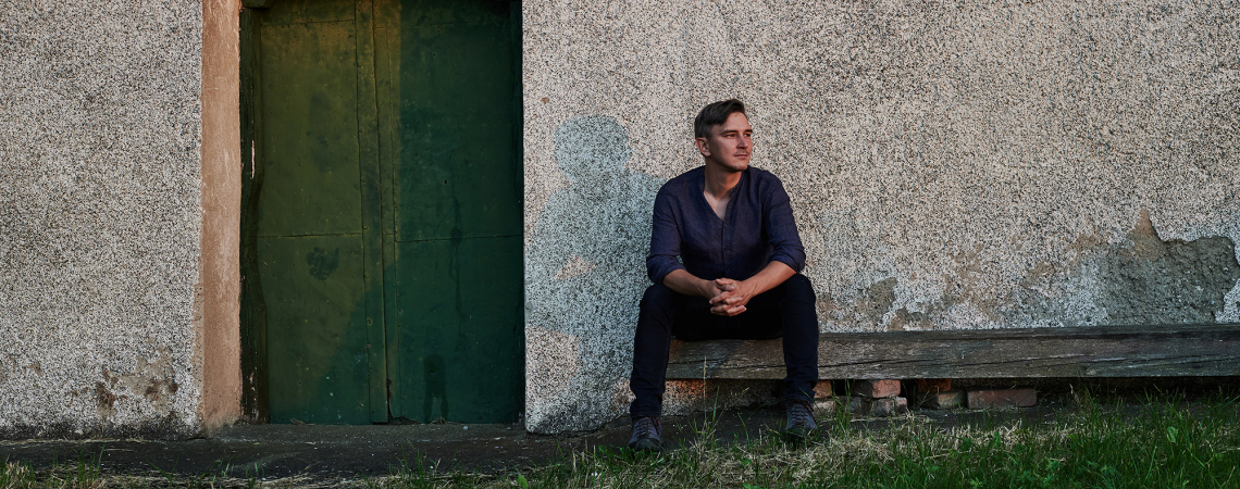 Mann sitzt auf Holzbank vor grüner Tür und rustikaler Hauswand.