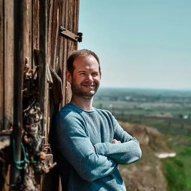 Mann lehnt sich lächelnd an Holzwand vor landschaftlichem Hintergrund.