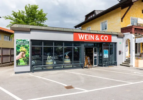 Kleiner Laden mit rotem Schild und großem Fenster, daneben Werbeplakat mit Traubenmotiv.