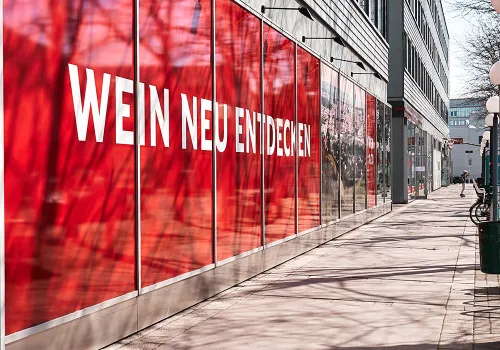 Rotes Schaufenster mit modernem Schriftzug an einem Bürogebäude entlang eines Gehwegs.
