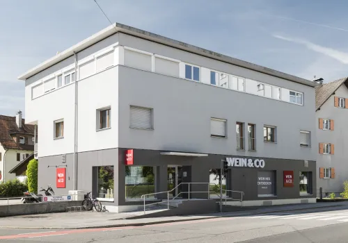 Modernes Eckgebäude mit großen Fenstern und markanter grauer Fassade an einer Straßenkreuzung.