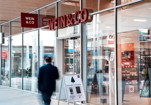 Moderner Weinladen mit großen Glasfenstern und rotem Schild; Person im Anzug läuft vorbei.