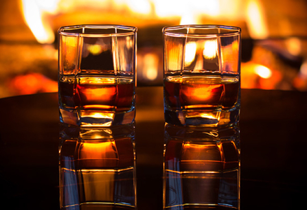Zwei Whiskygläser auf Tisch, reflektiert vor einem warmen Kaminfeuer.