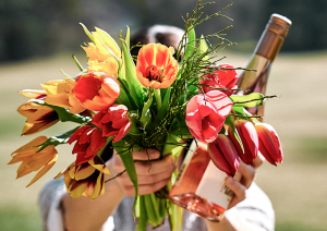 Bunter Tulpenstrauß und Roséweinflasche in Händen, mit unscharfem Naturhintergrund.