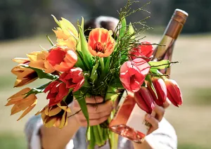 Bunter Tulpenstrauß und Roséweinflasche in Händen, mit unscharfem Naturhintergrund.