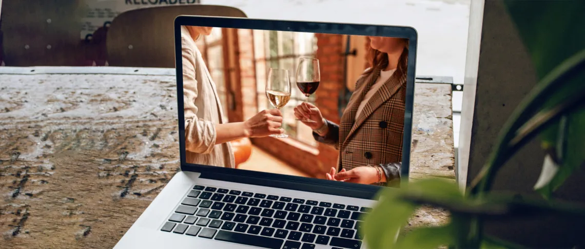 Laptop auf Holzfläche zeigt zwei Personen beim Anstoßen mit Wein.